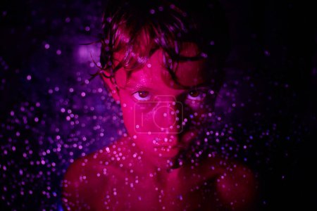 Foto de A través de la vista de la pared de cristal de niño irritado con el pelo mojado y el torso desnudo mirando a la cámara en luz bokeh púrpura - Imagen libre de derechos