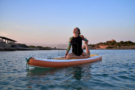 Photo for Full length of young female athlete in swimsuit stretching leg in Ardha Kapotasana pose while sitting on paddleboard in rippling sea on sunset - Royalty Free Image