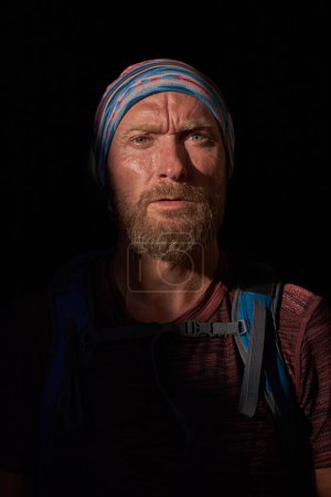 Photo for Pensive bearded traveling man in bandana with backpack frowning and looking at camera while standing in shadow with sunlight - Royalty Free Image