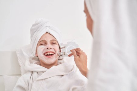 Téléchargez les photos : Femme avec serviette sur la tête appliquer un masque hydratant avec brosse sur le visage de l'enfant excité en peignoir relaxant pendant la séance de spa avec les yeux fermés - en image libre de droit