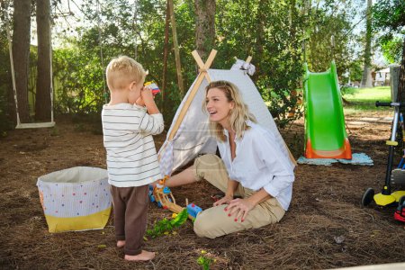 Téléchargez les photos : Corps complet de mignon garçon et mère en vêtements décontractés jouant avec des jouets près de wigwam dans un parc vert avec des enfants glissent - en image libre de droit