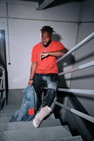 Photo for Full length of confident African American male model with dreadlocks wearing trendy outfit looking at camera and standing on staircase - Royalty Free Image