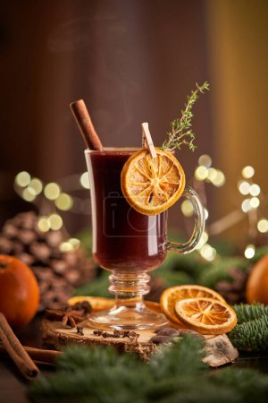 Foto de Copa de vino caliente alcohólico servido en la mesa con naranjas y ramas de coníferas durante la celebración de Navidad en la habitación con guirnalda brillante - Imagen libre de derechos