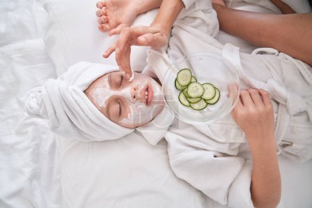 Photo for From above of crop anonymous mother applying moisturizing facial mask on kid with towel on head relaxing on comfortable bed with closed eyes and holding plate with cucumber slices - Royalty Free Image