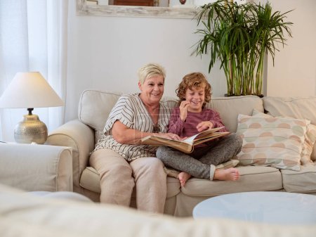Téléchargez les photos : Joyeux grand-mère aînée et petit garçon lecture ouvert livre photo ensemble et amusez-vous tout en passant du temps dans le salon confortable - en image libre de droit