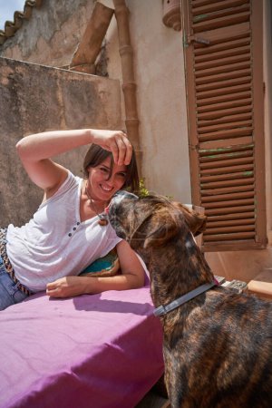 Téléchargez les photos : Femme souriante en vêtements décontractés s'amusant tout en étant allongé sur un banc avec matelas rose et en jouant avec le chien sur la terrasse de la maison en pierre le jour ensoleillé d'été - en image libre de droit