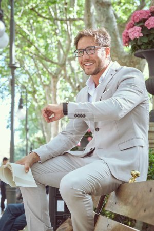 Téléchargez les photos : Corps complet de l'entrepreneur masculin ponctuel dans des lunettes détournant les yeux tout en vérifiant l'heure et en attendant un rendez-vous d'affaires dans le parc avec livre le jour ensoleillé - en image libre de droit
