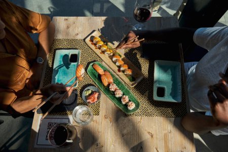 Photo for Top view of crop anonymous male in casual clothes sitting at wooden table with glass of wine and eating asian food for dinner together on sunny day - Royalty Free Image