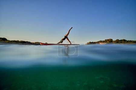 Téléchargez les photos : Athlète féminine anonyme en maillot de bain pratiquant le yoga sur la planche SUP et prenant la pose Adho Mukha Svanasana en mer au coucher du soleil - en image libre de droit