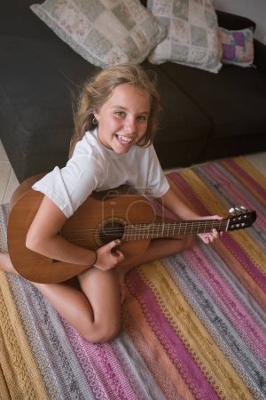 Téléchargez les photos : D'en haut de talentueuse fille souriant et regardant la caméra tout en jouant de la chanson à la guitare acoustique dans le salon à la maison - en image libre de droit