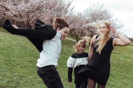 Téléchargez les photos : Femme heureuse et les enfants en tenues noires debout sur l'herbe verte près de la cerise en fleurs - en image libre de droit