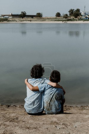 Téléchargez les photos : Vue arrière de garçons méconnaissables en uniforme scolaire sale et les cheveux bouclés assis sur la rive du lac avec les mains sur l'épaule tout en profitant de la journée d'été - en image libre de droit