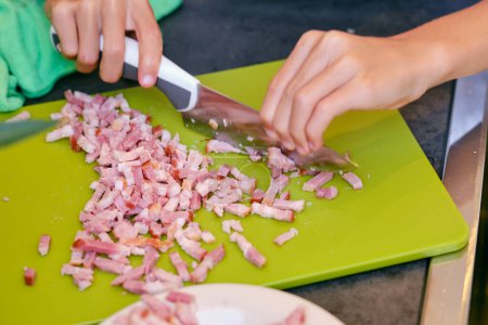 Photo for Crop anonymous person with knife cutting bacon on chopping board during cooking process at home - Royalty Free Image