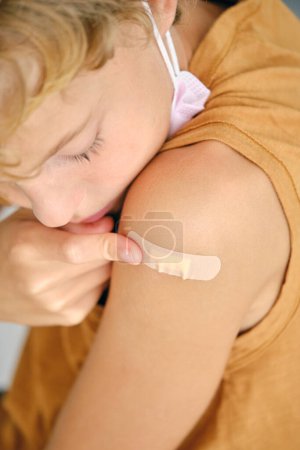 Téléchargez les photos : Cultivez l'enfant avec un masque stérile en appliquant un patch médical sur l'épaule pendant la pandémie de COVID 19 - en image libre de droit