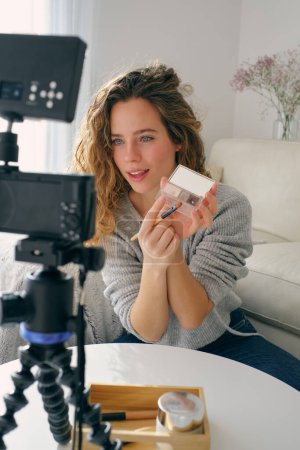 Photo for Young female blogger sitting at table with camera and shooting video while reviewing cosmetics in light room at home - Royalty Free Image