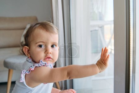Photo for Side view of adorable baby girl with big brown eyes looking at camera while leaning on window in living room at home - Royalty Free Image