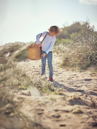 Téléchargez les photos : Corps complet de garçon préadolescent avec sac en osier ramassant les déchets en plastique et les ordures avec ramasse-ordures sur un sol sablonneux dans une journée ensoleillée - en image libre de droit