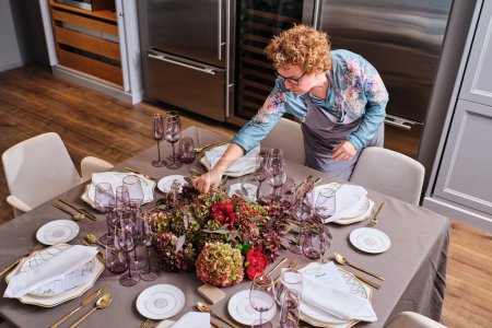Photo for From above of curly haired woman in eyeglasses and apron standing near table with served plates and glasses while making decoration with flowers for event - Royalty Free Image