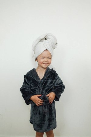 Foto de Encantadora niña feliz con albornoz gris y toalla blanca en la cabeza después del baño de pie contra la pared blanca en casa - Imagen libre de derechos