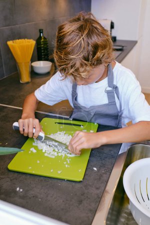 Photo for Concentrated kid in apron chopping raw onion with knife while cooking at table in house - Royalty Free Image