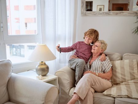 Téléchargez les photos : Mature grand-mère et garçon assis sur le canapé dans des vêtements décontractés étreignant et prenant selfie - en image libre de droit