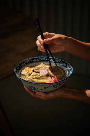 Photo for Crop anonymous female with chopsticks eating yummy ramen soup with noodles and meat in bowl against dark background - Royalty Free Image