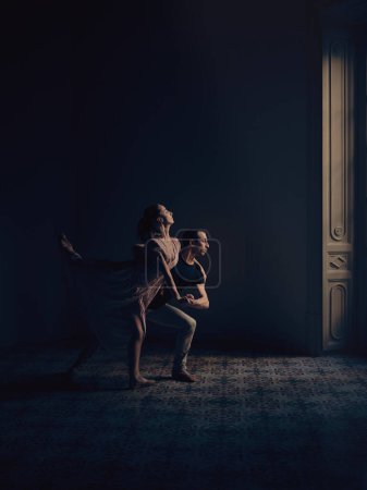 Foto de Vista lateral de cuerpo completo del talentoso hombre y mujer realizando movimiento de ballet mientras bailan juntos en estudio oscuro con columna - Imagen libre de derechos