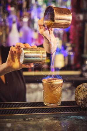 Téléchargez les photos : Cultiver méconnaissable femelle barman verser ingrédient dans un cocktail flamboyant debout au comptoir dans le bar moderne - en image libre de droit