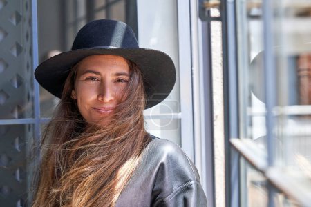 Téléchargez les photos : Portrait de jeune femme élégante en chapeau regardant la caméra tout en se tenant près de la fenêtre - en image libre de droit