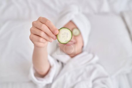 Photo for From above of child wearing bathrobe and towel turban lying on white bed and demonstrating cucumber slice during spa procedures in bedroom at home - Royalty Free Image
