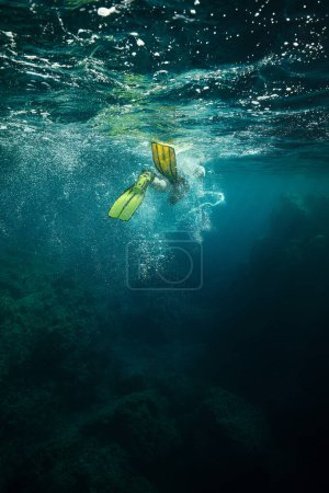Téléchargez les photos : Homme anonyme en palmes jaunes nageant sous l'eau de mer sombre avec mousse et bulles près de la surface et le fond avec des coraux - en image libre de droit
