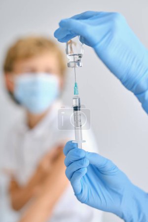 Foto de Cultivo médico irreconocible en guantes estériles con inyector de llenado de frasco pequeño con la vacuna COVID 19 contra el paciente en la clínica - Imagen libre de derechos