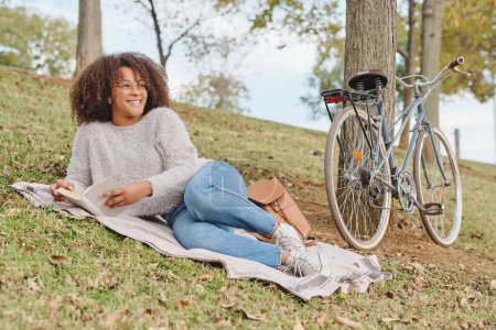 Foto de Vista lateral de la alegre mujer afroamericana en ropa casual sonriendo y mirando hacia otro lado mientras descansa sobre una manta cerca de la bicicleta y leyendo una interesante historia en el parque verde - Imagen libre de derechos