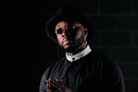 Téléchargez les photos : Homme afro-américain confiant en tenue noire tendance avec des accessoires debout contre un mur de briques dans une pièce sombre - en image libre de droit