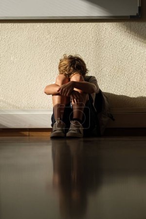 Téléchargez les photos : Triste écolier solitaire assis sur le sol dans la salle de classe à l'école et embrassant genoux - en image libre de droit