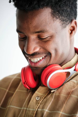 Foto de Joven hombre afroamericano en camisa a cuadros con auriculares contemporáneos mirando hacia abajo con sonrisa dentada - Imagen libre de derechos