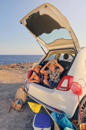 Téléchargez les photos : Photo verticale de deux enfants assis dans le coffre de la voiture faisant des gestes de blague avec des paniers d'objets pour la plage et une glacière portable à côté de la voiture avec la mer en arrière-plan - en image libre de droit
