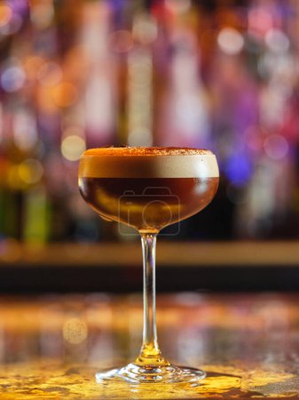 Téléchargez les photos : Verre plein de cocktail expresso brun alcoolisé avec mousse blanche servi dans un bar avec des bouteilles sur fond flou avec bokeh - en image libre de droit