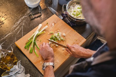 Photo for From above of crop anonymous male cook cutting fresh green onion with knife on chopping board at counter in light kitchen - Royalty Free Image