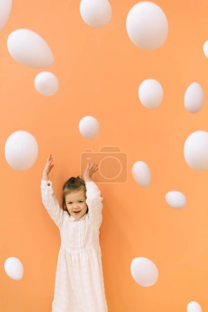 Foto de Adorable niña en vestido blanco de pie cerca de la pared naranja y lanzando huevos de Pascua - Imagen libre de derechos