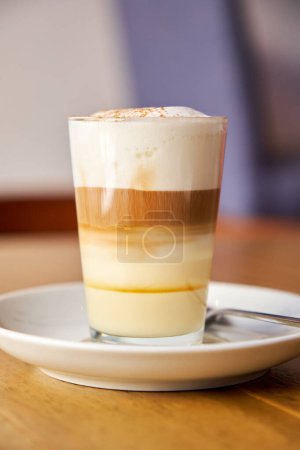 Foto de Foto vertical de un café con leche servido en un vaso de cristal encima de un plato de porcelana sobre una superficie de madera - Imagen libre de derechos