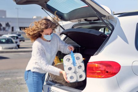Photo for Attractive young female wearing casual outfit and face mask placing grocery bags and toilet paper packs in car trunk parked on sunny parking near supermarket - Royalty Free Image