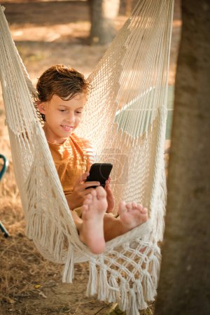 Photo for Glad barefoot boy smiling and browsing cellphone while chilling in hammock in countryside - Royalty Free Image