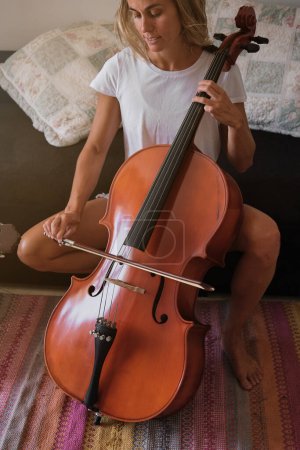 Foto de Talentosa mujer músico con violín sentado en el sofá y tocando melodía en violonchelo en la sala de estar en casa en el día de fin de semana - Imagen libre de derechos