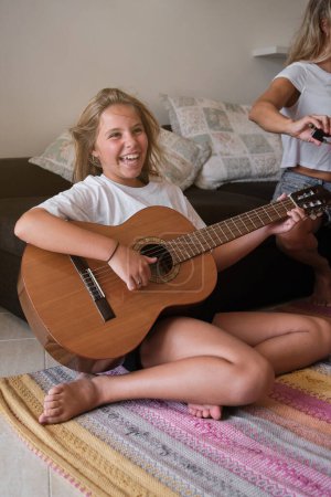 Photo for Full length of laughing girl in casual clothes having fun while sitting on carpet near sofa in living room and playing acoustic guitar on weekend day - Royalty Free Image