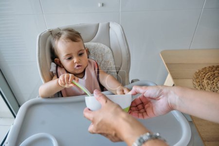Photo for Adorable little girl in bib sitting on white high chair and eating with spoon while unrecognizable woman helping her and holding bowl - Royalty Free Image