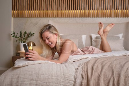 Photo for Side view of young barefoot woman in pink nightie having fun while reading text messages on phone and relaxing in cozy bedroom at home - Royalty Free Image