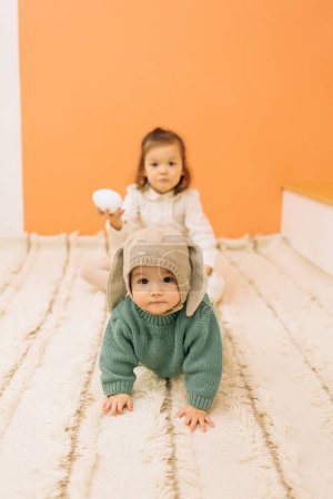 Photo for Full body of adorable baby girl and toddler sister with egg sitting on carpet floor in room with orange wall - Royalty Free Image
