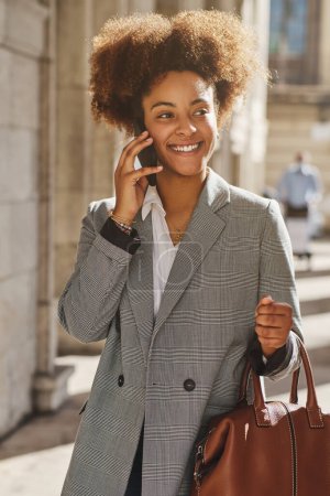 Téléchargez les photos : Femme d'affaires ethnique positive avec coiffure afro regardant loin tout en se tenant dans la rue avec sac et parlant sur téléphone portable en été - en image libre de droit