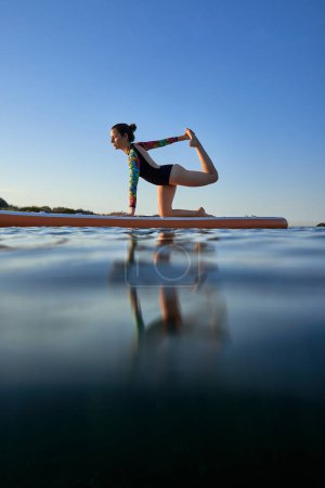 Téléchargez les photos : Pleine longueur de sportive prenant la pose de Vyaghrasana et étirant la jambe sur le conseil SUP tout en pratiquant le yoga parmi l'eau de mer ondulante - en image libre de droit